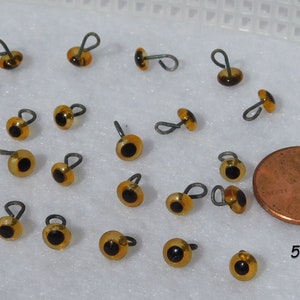 May include: A collection of 20 amber-colored plastic safety eyes with black pupils and a wire loop for sewing. The eyes are approximately 5mm in diameter and are shown next to a US penny for size comparison.