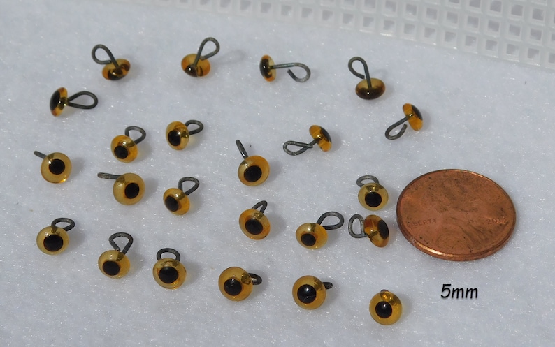 May include: A collection of 20 amber-colored plastic safety eyes with black pupils and a wire loop for sewing. The eyes are approximately 5mm in diameter and are shown next to a US penny for size comparison.