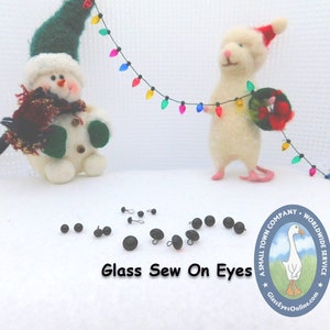 May include: A collection of black glass sew-on eyes for crafts. The eyes are arranged on a white surface with a snowman and a mouse figurine in the background. The image also includes a logo for a company called "A Small Town Company - Worldwide Service" with the text "GlassEyesOnline.com".