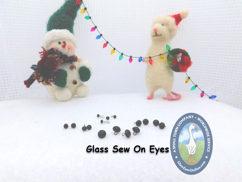 May include: A collection of black glass sew-on eyes for crafts. The eyes are arranged on a white surface with a snowman and a mouse figurine in the background. The image also includes a logo for a company called "A Small Town Company - Worldwide Service" with the text "GlassEyesOnline.com".