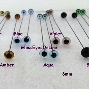 Op de afbeelding: Assortiment van 6 mm glazen ogen in verschillende kleuren, waaronder licht amber, donker amber, blauw, aqua, violet, groen en zwart. De ogen zijn gemonteerd op pinnen voor eenvoudig gebruik in knutselwerk.