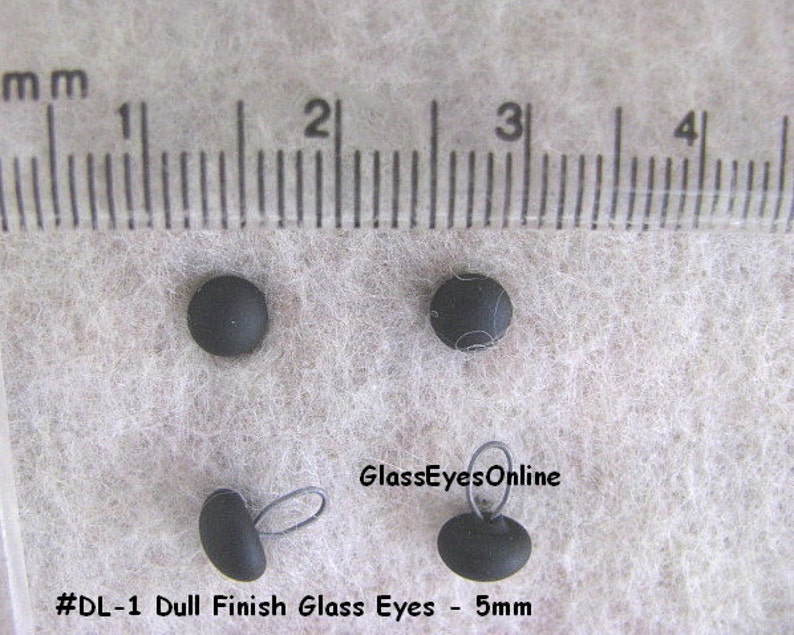 Op de afbeelding: Vier zwarte glazen ogen met een matte afwerking, met een diameter van 5 mm. De ogen worden getoond naast een liniaal ter referentie van de grootte. De tekst "#DL-1 Dull Finish Glass Eyes - 5mm" en "GlassEyesOnline" is zichtbaar in de afbeelding.