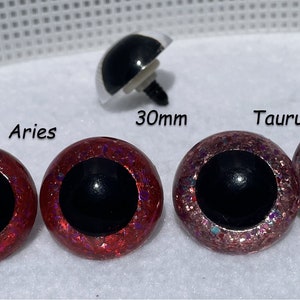 May include: Two pairs of 30mm safety eyes with black pupils. One pair is red with iridescent glitter, and the other pair is pink with iridescent glitter. The eyes are labeled "Aries" and "Taurus".