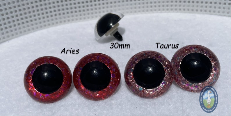 May include: Two pairs of 30mm safety eyes with black pupils. One pair is red with iridescent glitter, and the other pair is pink with iridescent glitter. The eyes are labeled "Aries" and "Taurus".
