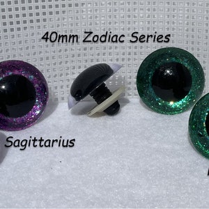 May include: Two pairs of 40mm glitter safety eyes in purple and green with black pupils. The eyes are labeled "Sagittarius" and "Pisces" and are part of a Zodiac series.