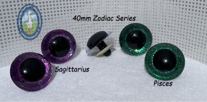 May include: Two pairs of 40mm glitter safety eyes in purple and green with black pupils. The eyes are labeled "Sagittarius" and "Pisces" and are part of a Zodiac series.