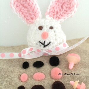 May include: A white crocheted bunny with pink ears and brown eyes. The bunny is surrounded by a variety of pink and brown pom-poms and two sets of plastic safety eyes.