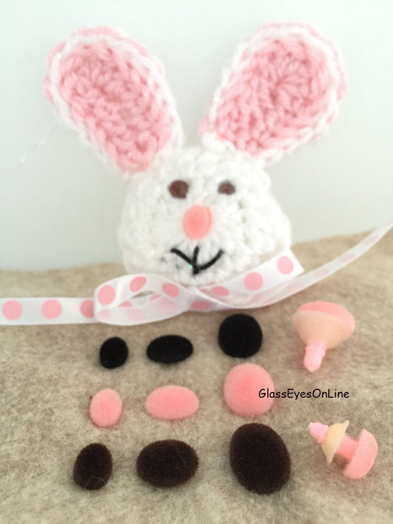 May include: A white crocheted bunny with pink ears and brown eyes. The bunny is surrounded by a variety of pink and brown pom-poms and two sets of plastic safety eyes.
