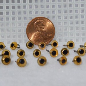 May include: A close-up of a group of 20 amber-colored plastic safety eyes with black pupils and silver metal shanks. The eyes are arranged in a random pattern on a white surface. A penny is included for size comparison.