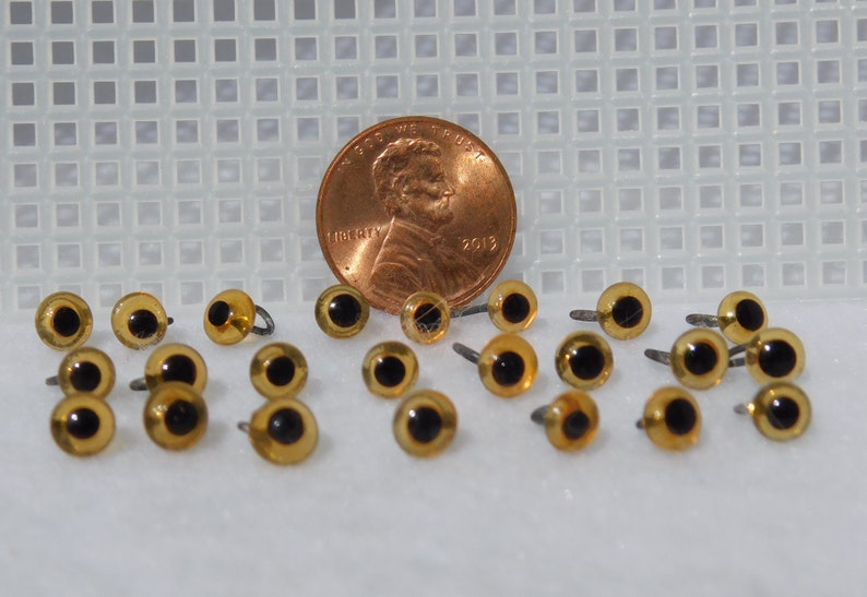 May include: A close-up of a group of 20 amber-colored plastic safety eyes with black pupils and silver metal shanks. The eyes are arranged in a random pattern on a white surface. A penny is included for size comparison.