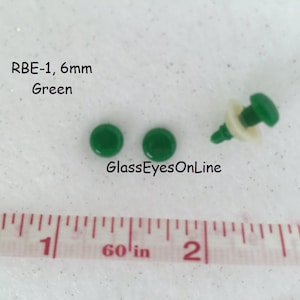 K&ouml;nnte beinhalten: Zwei gr&uuml;ne 6 mm Kunststoffaugen mit wei&szlig;er R&uuml;ckseite. Die Augen befinden sich auf einer wei&szlig;en Oberfl&auml;che mit einem Lineal, das 1 Zoll anzeigt. Der Text "RBE-1, 6 mm Green" befindet sich &uuml;ber den Augen. Der Text "GlassEyesOnLine" befindet sich unter den Augen.