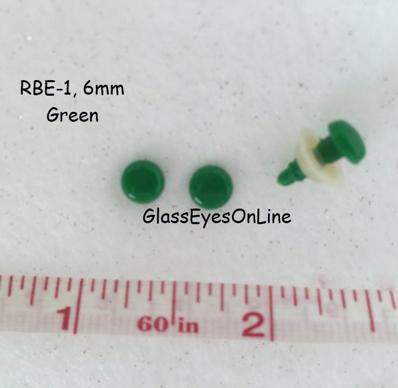 K&ouml;nnte beinhalten: Zwei gr&uuml;ne 6 mm Kunststoffaugen mit wei&szlig;er R&uuml;ckseite. Die Augen befinden sich auf einer wei&szlig;en Oberfl&auml;che mit einem Lineal, das 1 Zoll anzeigt. Der Text "RBE-1, 6 mm Green" befindet sich &uuml;ber den Augen. Der Text "GlassEyesOnLine" befindet sich unter den Augen.