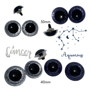 May include: Two pairs of plastic safety eyes with black pupils and silver and blue glitter. The eyes are 30mm and 40mm in diameter. The text "Cancer" and "Aquarius" are written below the eyes.