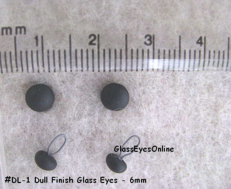Op de afbeelding: Vier zwarte glazen ogen met een matte afwerking, met een diameter van 6 mm. Twee van de ogen hebben een zwarte lus om te naaien.