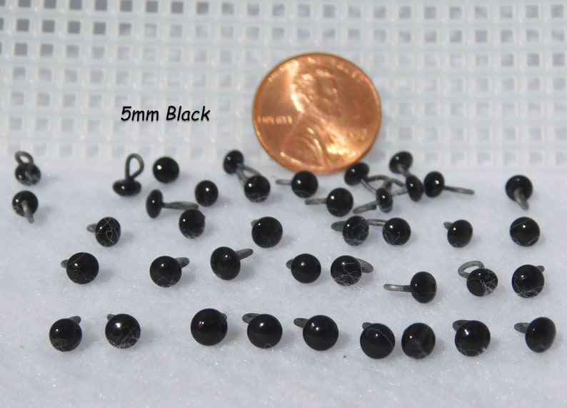 May include: A close-up of 5mm black plastic safety eyes with a metal loop for sewing. The eyes are shown next to a US penny for size comparison. 5mm Black.