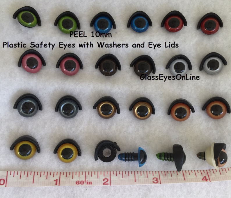 Puede incluir: Surtido de ojos de seguridad de pl&aacute;stico de 10 mm con arandelas y p&aacute;rpados en varios colores, incluidos verde, azul, rojo, rosa, marr&oacute;n, gris, dorado y negro. Los ojos est&aacute;n dispuestos en filas sobre una superficie blanca con una regla para la escala. El texto "PEEL 10mm" y "Plastic Safety Eyes with Washers and Eye Lids" es visible en la parte superior de la imagen. El texto "GlassEyesOnLine" es visible en la parte inferior de la imagen.
