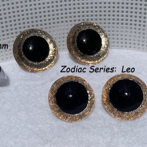 May include: Four black and gold glitter safety eyes for crafts. Two eyes are 40mm and two are 34mm. The eyes have a gold glitter rim and a black center. The text "Zodiac Series: Leo" is visible in the image.