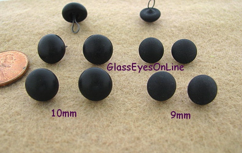 Op de afbeelding: Assortiment van zwarte plastic knoppen in twee maten, 9 mm en 10 mm. De knoppen zijn rond en hebben een gladde, matte afwerking. GlassEyesOnLine is op het oppervlak gedrukt.