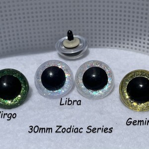 May include: Set of six 30mm glitter safety eyes for plush making. The eyes are green, white, and gold with black pupils. The text "30mm Zodiac Series" is printed below the eyes. The eyes are labeled "Virgo", "Libra", and "Gemini".