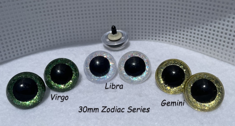 May include: Set of six 30mm glitter safety eyes for plush making. The eyes are green, white, and gold with black pupils. The text "30mm Zodiac Series" is printed below the eyes. The eyes are labeled "Virgo", "Libra", and "Gemini".