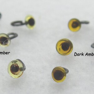 May include: A close-up of two different colors of plastic safety eyes for crafts. The eyes are amber in color, with a black pupil. The eyes are on a white surface. The text "Light Amber" and "Dark Amber" are written below the eyes.