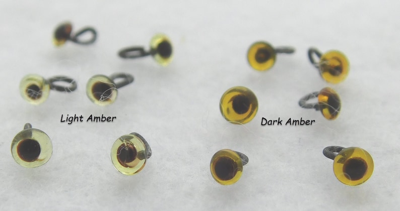 May include: A close-up of two different colors of plastic safety eyes for crafts. The eyes are amber in color, with a black pupil. The eyes are on a white surface. The text "Light Amber" and "Dark Amber" are written below the eyes.