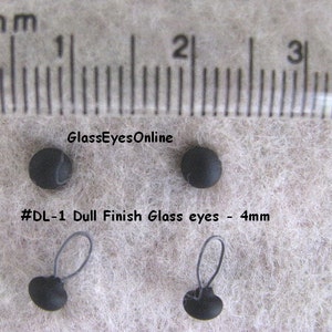 Op de afbeelding: Vier zwarte glazen ogen met een matte afwerking, met een diameter van 4 mm. Twee van de ogen hebben een zwarte lus aan de achterkant. De ogen worden getoond op een wit, pluche oppervlak met een liniaal op de achtergrond.