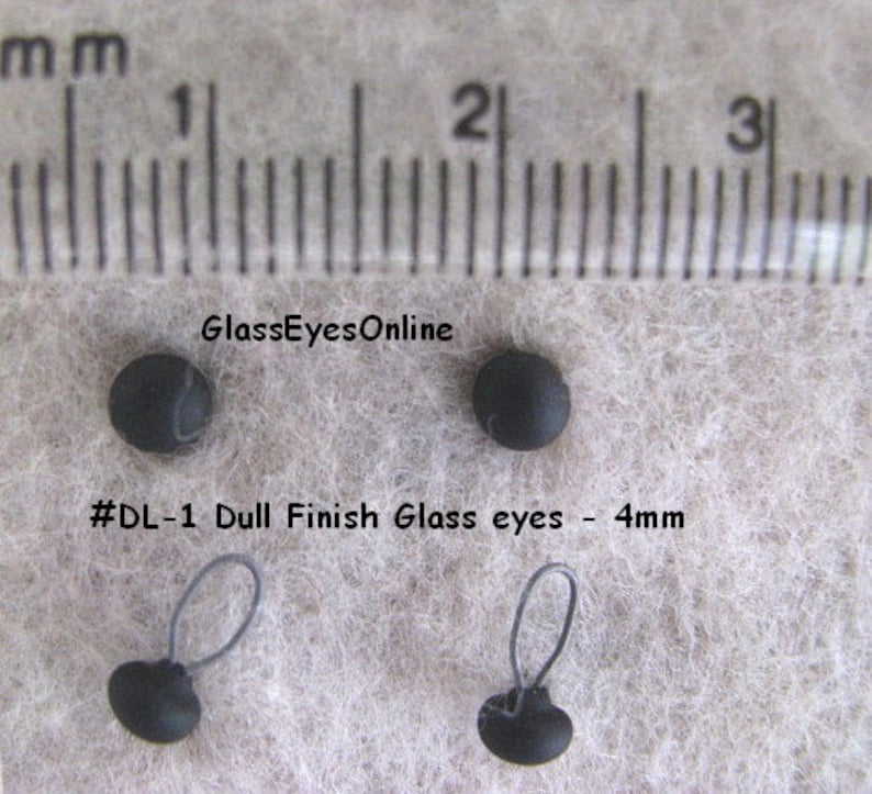 Op de afbeelding: Vier zwarte glazen ogen met een matte afwerking, met een diameter van 4 mm. Twee van de ogen hebben een zwarte lus aan de achterkant. De ogen worden getoond op een wit, pluche oppervlak met een liniaal op de achtergrond.