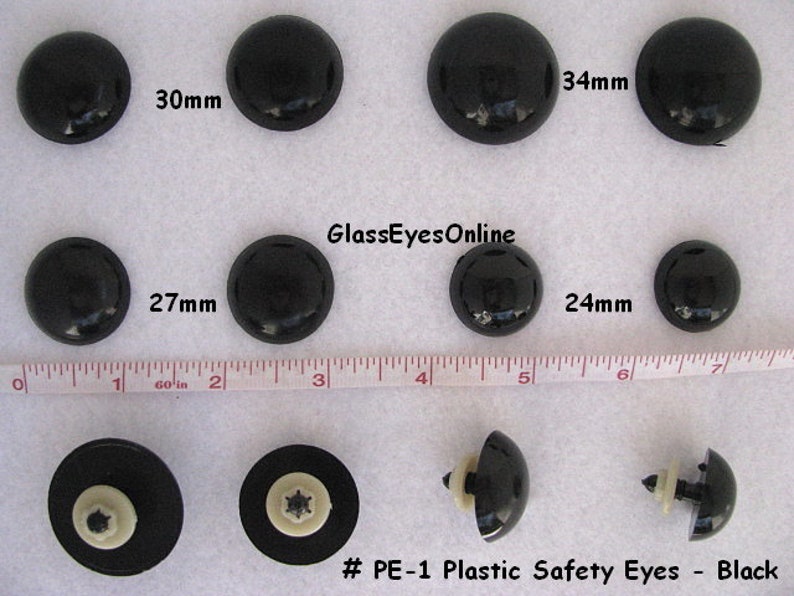 May include: Assortment of black plastic safety eyes in various sizes, ranging from 24mm to 34mm. The eyes are labelled with their size in millimetres. The text "#PE-1 Plastic Safety Eyes - Black" is displayed at the bottom of the image.