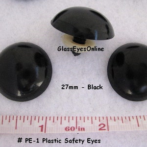 May include: Three black plastic safety eyes, each measuring 27mm in diameter. The eyes have a flat back and a raised dome shape. The text "GlassEyesOnline" and "27mm - Black" are visible below the eyes. A ruler is visible at the bottom of the image with the text "# PE-1 Plastic Safety Eyes" and "60' in".