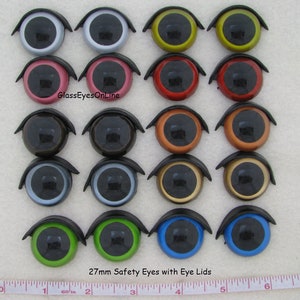 Puede incluir: Veintisiete ojos de seguridad de 27 mm con p&aacute;rpados en varios colores, incluidos negro, blanco, rojo, verde, azul, marr&oacute;n, dorado, plateado y naranja. Los ojos est&aacute;n dispuestos en tres filas de nueve, con una regla en la parte inferior de la imagen para la escala. GlassEyesOnline.