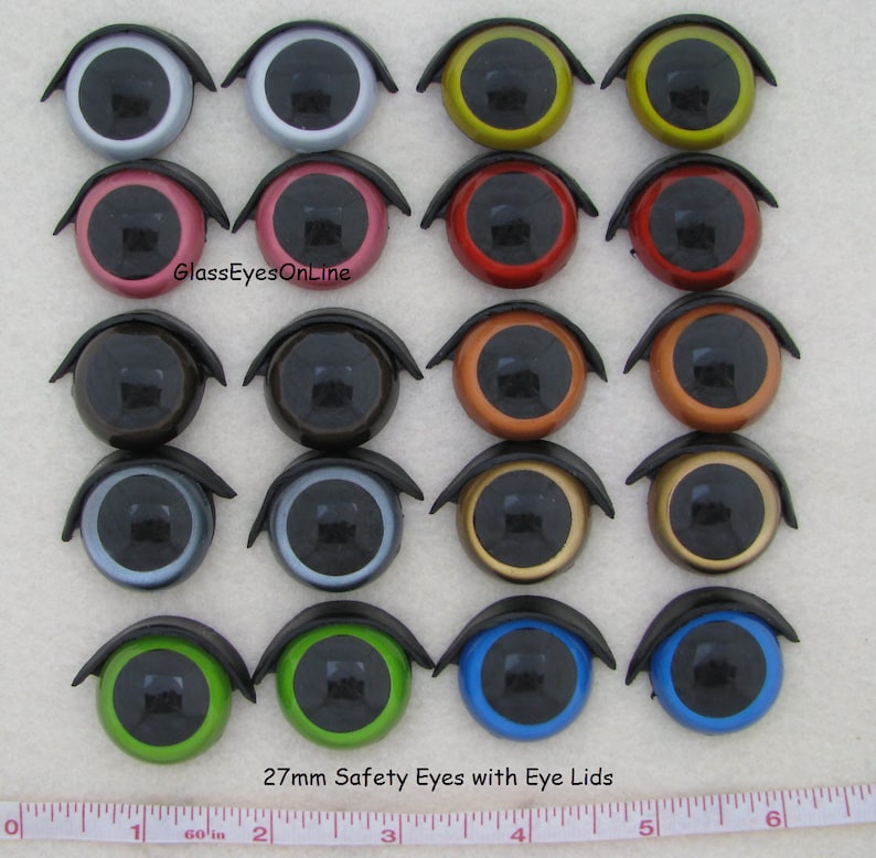Puede incluir: Veintisiete ojos de seguridad de 27 mm con p&aacute;rpados en varios colores, incluidos negro, blanco, rojo, verde, azul, marr&oacute;n, dorado, plateado y naranja. Los ojos est&aacute;n dispuestos en tres filas de nueve, con una regla en la parte inferior de la imagen para la escala. GlassEyesOnline.