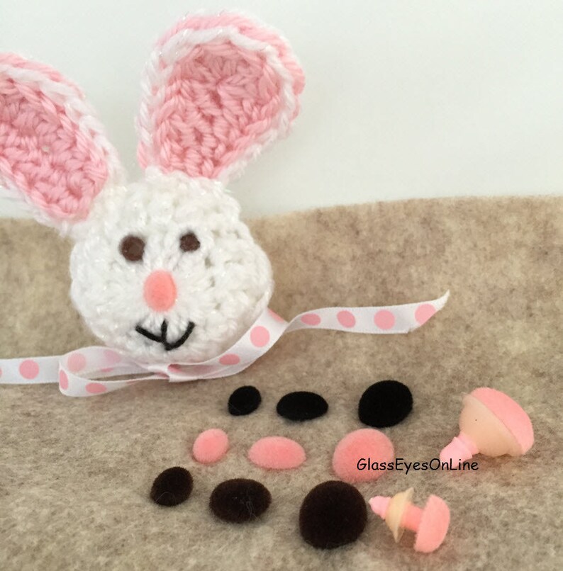 May include: A white crocheted bunny head with pink ears and brown eyes.  The bunny is surrounded by black and pink felt circles and two plastic safety eyes with pink lids.