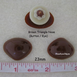 Pode incluir: Dois olhos de vidro de forma triangular marrom, cada um com um di&acirc;metro de 23 mm, com um nariz branco em forma de bot&atilde;o no centro. Os olhos est&atilde;o sobre uma superf&iacute;cie branca com uma r&eacute;gua para a escala.