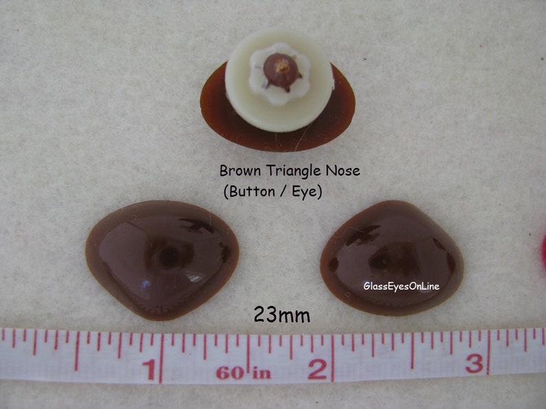 Pode incluir: Dois olhos de vidro de forma triangular marrom, cada um com um di&acirc;metro de 23 mm, com um nariz branco em forma de bot&atilde;o no centro. Os olhos est&atilde;o sobre uma superf&iacute;cie branca com uma r&eacute;gua para a escala.