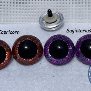 May include: Two pairs of plastic safety eyes for crafts. The eyes are 12mm in diameter and have a black pupil. One pair is a copper color with glitter, labeled "Capricorn". The other pair is purple with glitter, labeled "Sagittarius".