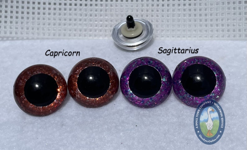 May include: Two pairs of plastic safety eyes for crafts. The eyes are 12mm in diameter and have a black pupil. One pair is a copper color with glitter, labeled "Capricorn". The other pair is purple with glitter, labeled "Sagittarius".