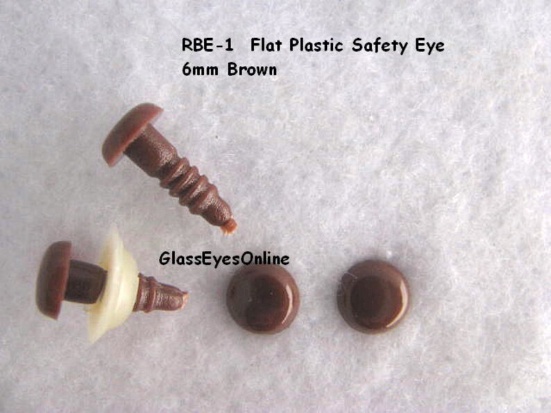 K&ouml;nnte beinhalten: Drei braune, flache Sicherheitsaugen aus Kunststoff, zwei davon flach und eines mit einer wei&szlig;en Unterlegscheibe. Die Augen haben einen Durchmesser von 6 mm. Der Text "RBE-1 Flat Plastic Safety Eye 6mm Brown" ist &uuml;ber den Augen sichtbar.