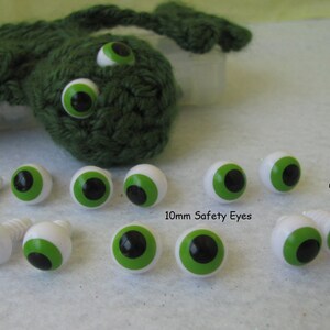 May include: A collection of 10mm green and black safety eyes with white plastic backs. The eyes are arranged in a row on a white surface. The text "GlassEyesOnLine" and "10mm Safety Eyes" are visible in the image.