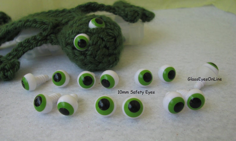 May include: A collection of 10mm green and black safety eyes with white plastic backs. The eyes are arranged in a row on a white surface. The text "GlassEyesOnLine" and "10mm Safety Eyes" are visible in the image.