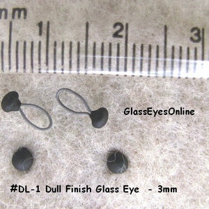 Op de afbeelding: Twee zwarte glazen ogen met een matte afwerking, elk 3 mm in diameter. De ogen zijn bevestigd aan zwarte lussen van touw. De afbeelding bevat een liniaal voor schaal en de tekst "#DL-1 Dull Finish Glass Eye - 3mm" en "GlassEyesOnline".