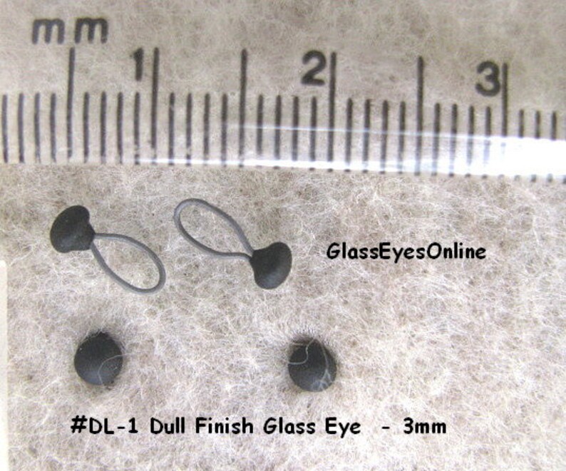 Op de afbeelding: Twee zwarte glazen ogen met een matte afwerking, elk 3 mm in diameter. De ogen zijn bevestigd aan zwarte lussen van touw. De afbeelding bevat een liniaal voor schaal en de tekst "#DL-1 Dull Finish Glass Eye - 3mm" en "GlassEyesOnline".