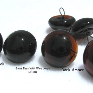 May include: Glass eyes with wire loops in black and dark amber colors. The eyes are round and have a black pupil. The text "Glass Eyes With Wire Loops LP-201" is printed below the eyes.
