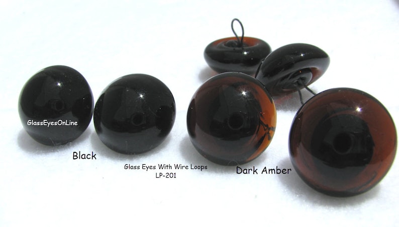 May include: Glass eyes with wire loops in black and dark amber colors. The eyes are round and have a black pupil. The text "Glass Eyes With Wire Loops LP-201" is printed below the eyes.