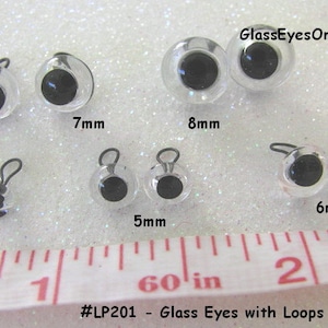May include: Assortment of clear glass eyes with black pupils and metal loops. The eyes are arranged in rows by size, with measurements in millimeters. The text "#LP201 - Glass Eyes with Loops - Clear" and "GlassEyesOnline" are visible in the image.