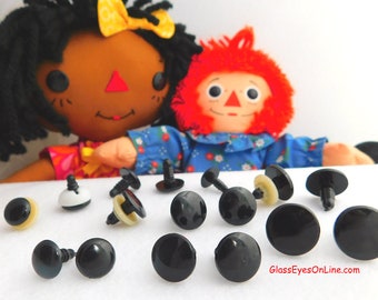 May include: A close-up of 12 black plastic safety eyes with a white washer and a screw back. The eyes are arranged in a semi-circle on a white surface. Two dolls are visible in the background.
