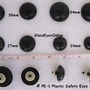 May include: Assortment of black plastic safety eyes in various sizes, ranging from 24mm to 34mm. The eyes are shown on a white surface with a ruler for size reference. The text "#PE-1 Plastic Safety Eyes - Black" is displayed below the eyes.
