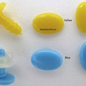 May include: Two oval-shaped glass eyes, one yellow and one blue, with a small plastic eye socket in the background. The text "GlassEyesOnline" is visible on the yellow eye.