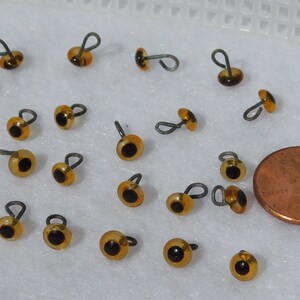 May include: A collection of 20 amber-colored plastic safety eyes with black pupils and a wire loop for attaching. The eyes are about the size of a US penny.