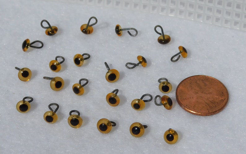 May include: A collection of 20 amber-colored plastic safety eyes with black pupils and a wire loop for attaching. The eyes are about the size of a US penny.